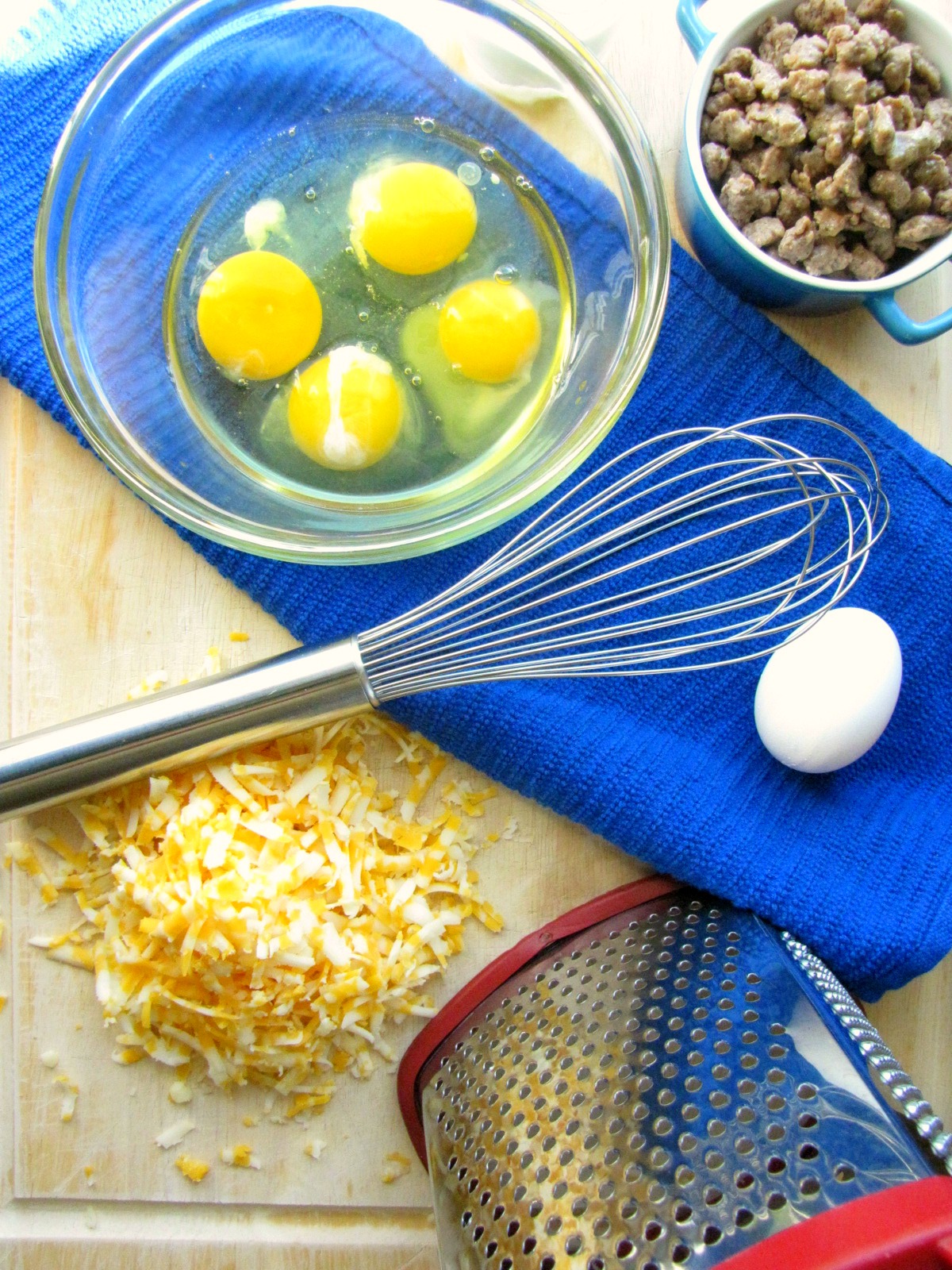 Four eggs in clear bowl, grated cheese, sausage with whisk 