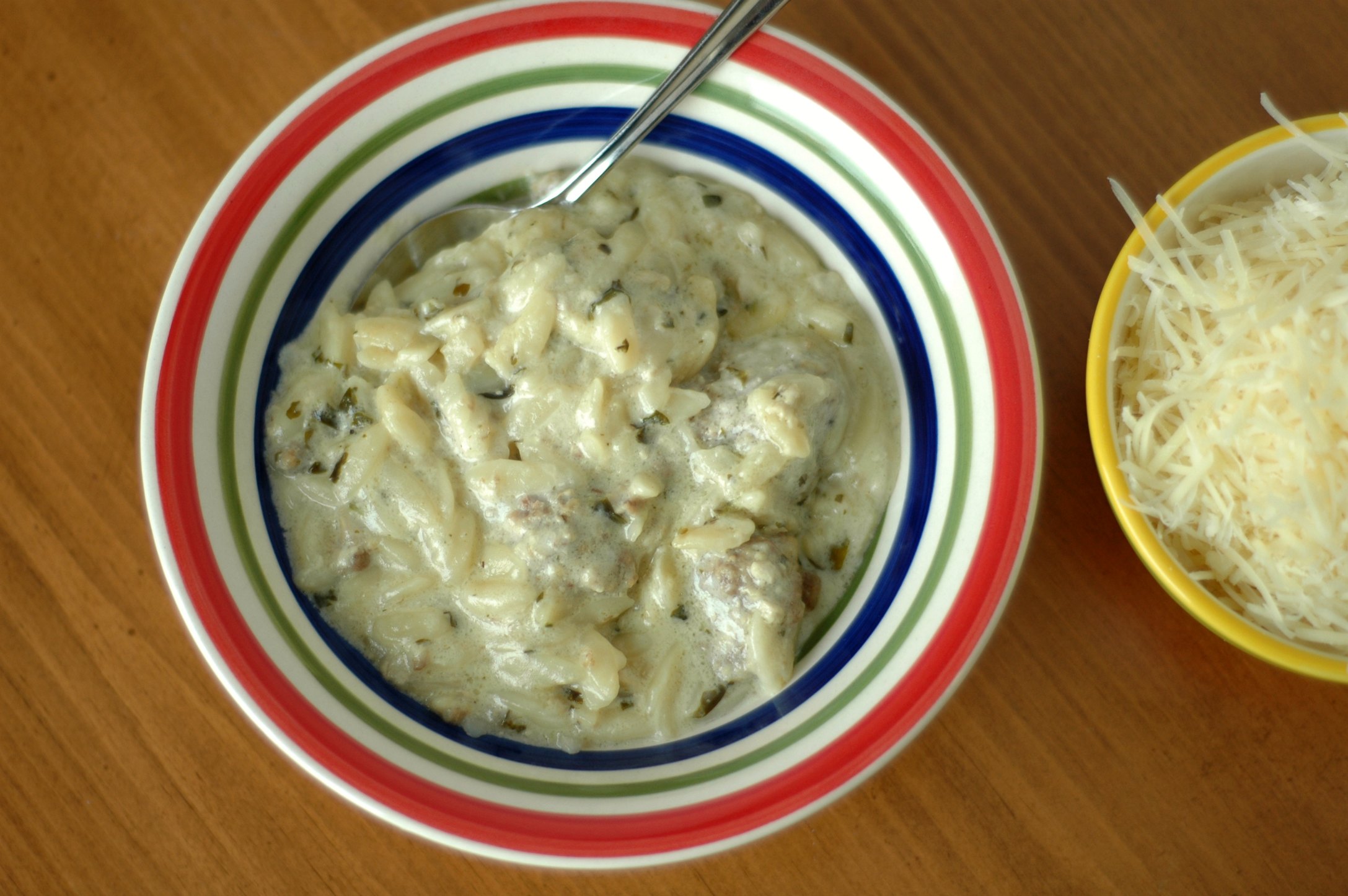Closeup of Creamy Meatball and Orzo Soup in white bowl with colored rim, bowl of shredded cheese