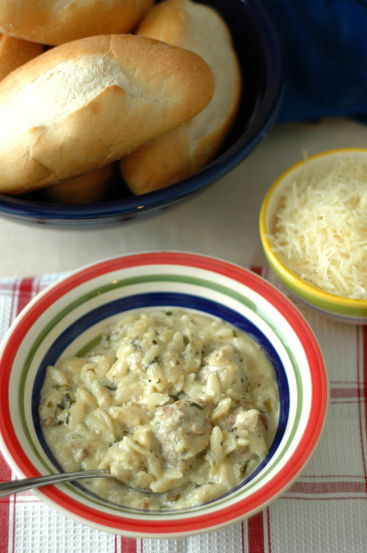 White bowl with colored rim of Creamy Meatball and Orzo Soup, baked roll and shredded cheese
