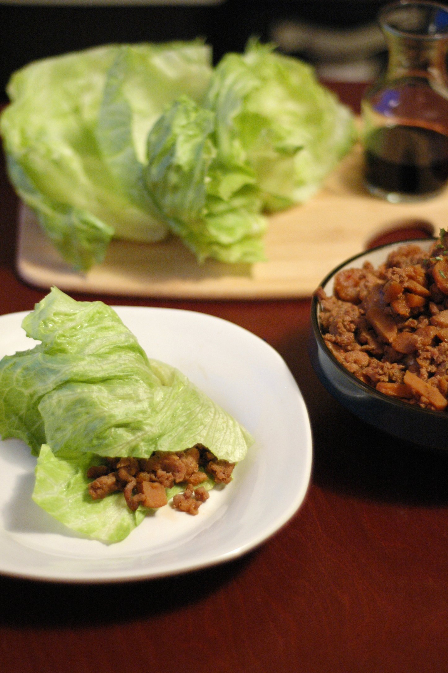 lettuce wrap on white plate, head of lettuce on cutting board and black bowl with pork mixture in background