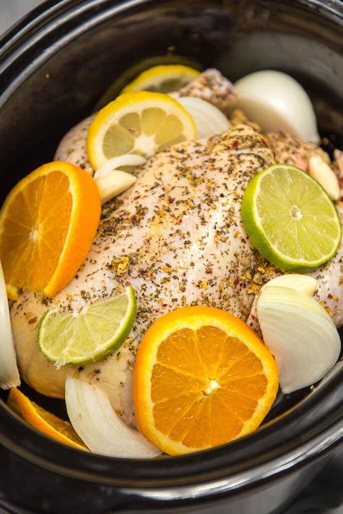 Whole chicken in black crock pot seasoned with herbs and covered on the sides with lemon and lime wedges, onions and garlic.