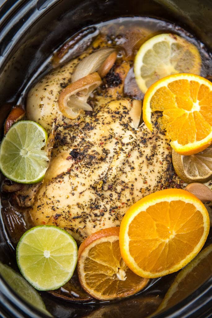 Cooked slow cooker whole chicken in black slow cooker covered in herbs with lemon and lime slices, onion and garlic surrounding it.