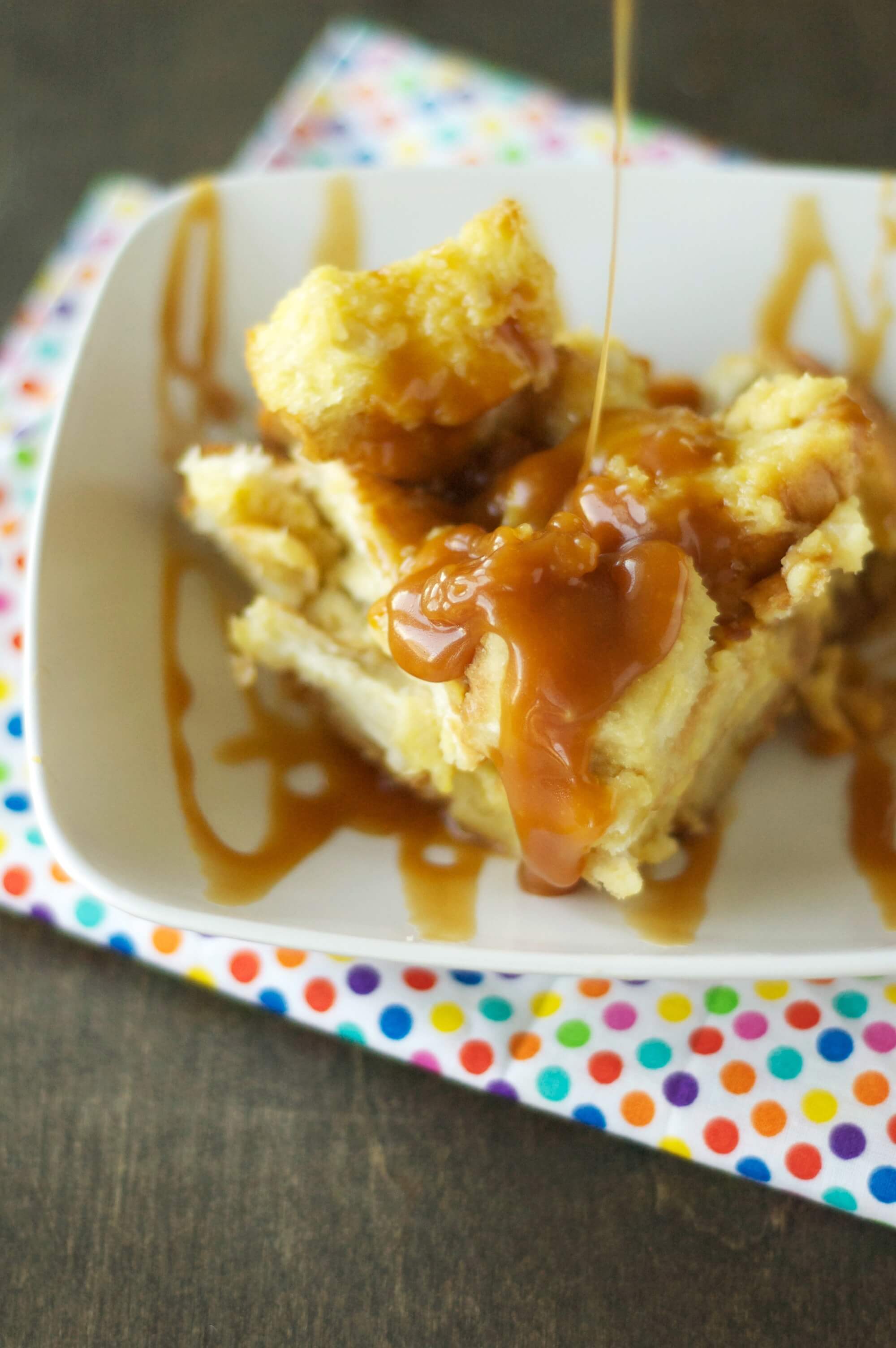 bread pudding being drizzled with salted caramel sauce in white bowl on polka dot napkin