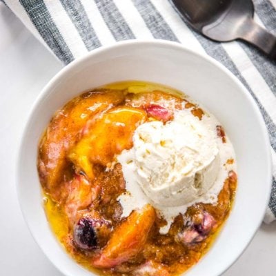 slow cooker peach cobbler with cherries topped with vanilla ice cream scoop in white bowl