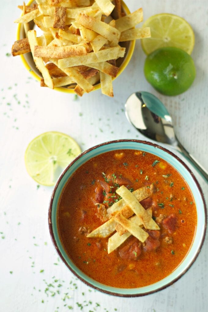Slow Cooker Beef Enchilada Soup in green bowl with bowl of tortilla strips, lime slices and spoon
