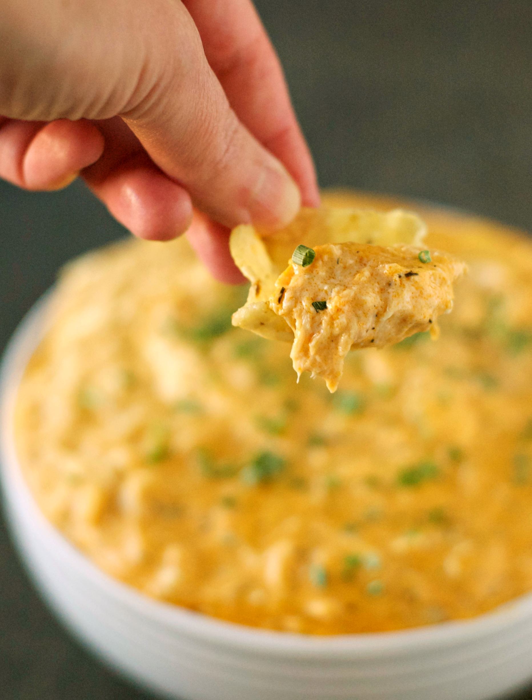 Slow Cooker Cheesy Buffalo Chicken Dip in white bowl with closeup of hand holding dip on chip