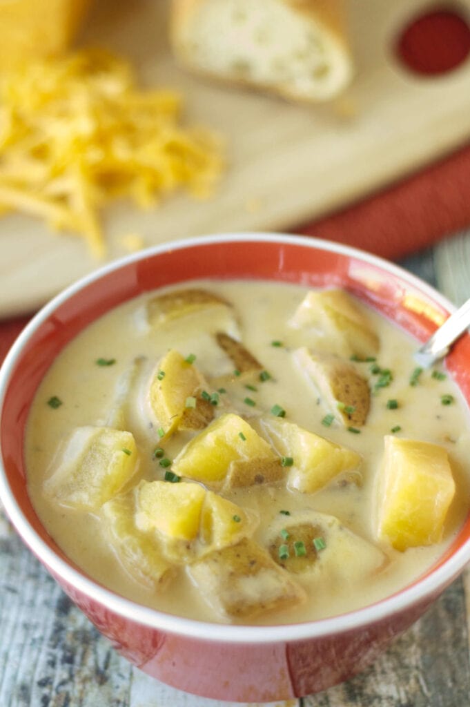 soup with potatoes in red bowl with spoon and board with bread and cheese in background