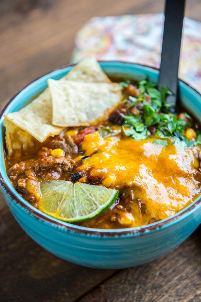 taco soup with tortilla chips, cheese, cilantro and lime wedge in blue bowl with black spoon