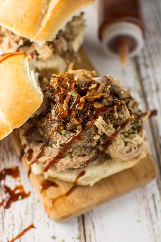 two pulled pork sandwiches on buns, one uncovered, with bbq sauce drizzled over on wood cutting board and bbq sauce bottle in background