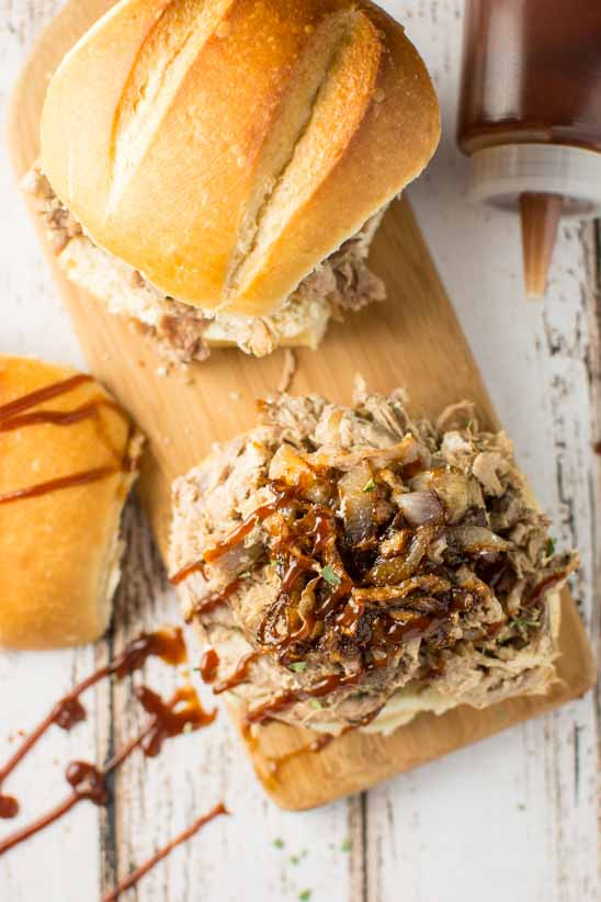 two pulled pork sandwiches on buns, one uncovered, with bbq sauce drizzled over on wood cutting board