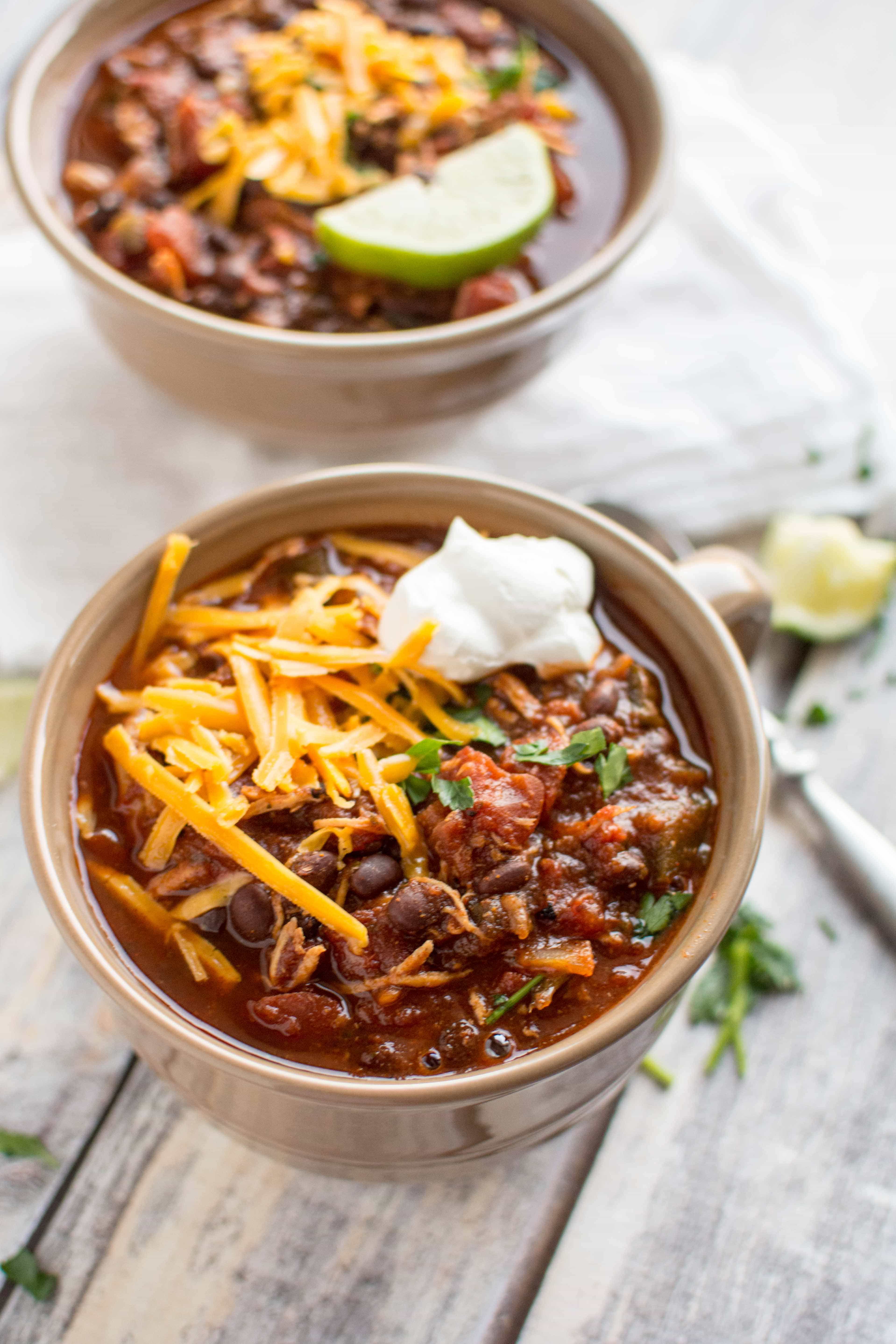 roasted tomato and ancho chicken chili in two tan serving bowls with cheese, lime and sour cream