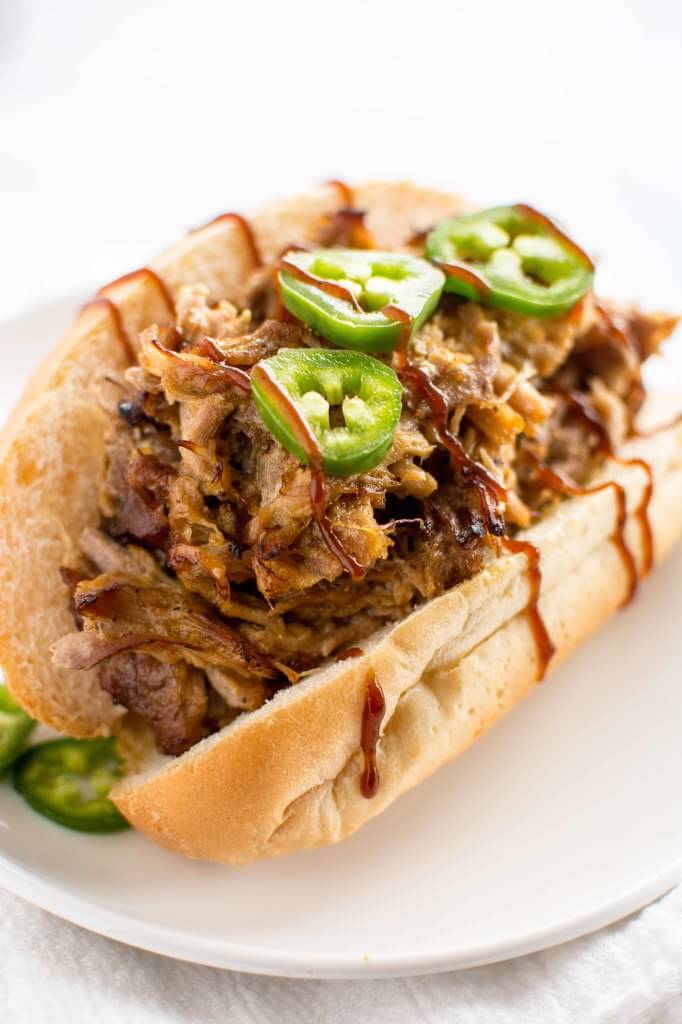 white plate with pulled pork on hoagie bun and 3 slices of jalapeno and drizzle of bbq sauce on top