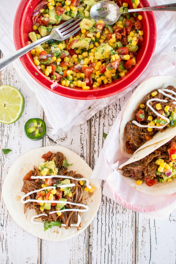 large red bowl with corn salsa with fork and spoon, one beef taco on table, and two shredded beef tacos in parchment lined basket with corn salsa on top