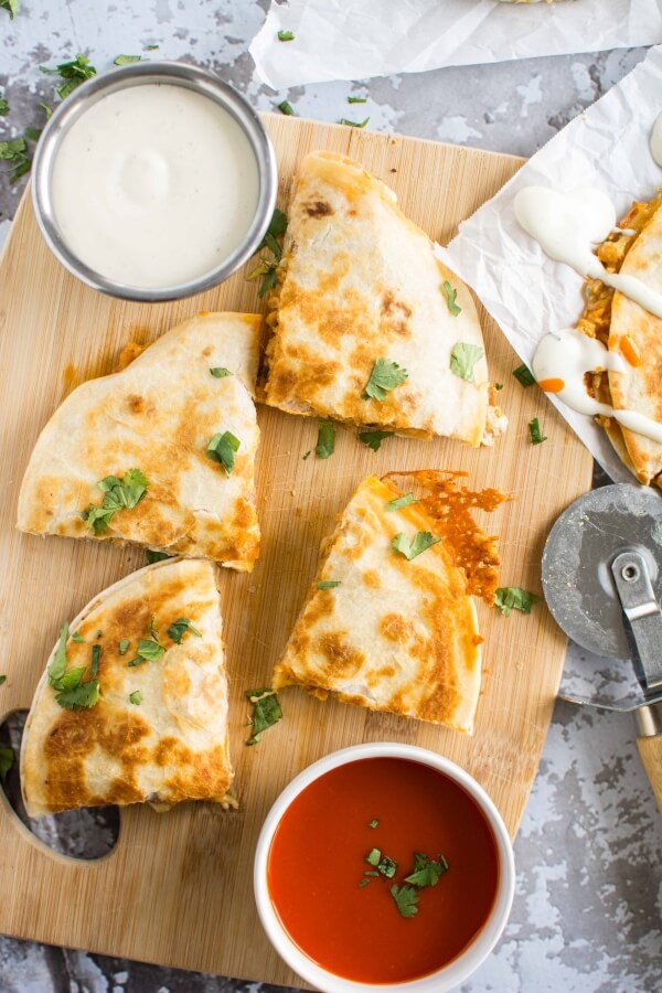 quesadilla cut in 4 pieces on wooden cutting board, with bowl of ranch and red sauce on side