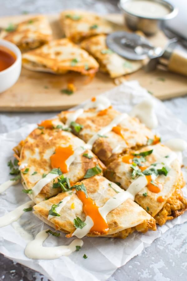 cut up quesadilla on parchment paper drizzled with ranch and buffalo sauce; cutting board with red and white sauce and four pieces of quesadilla 