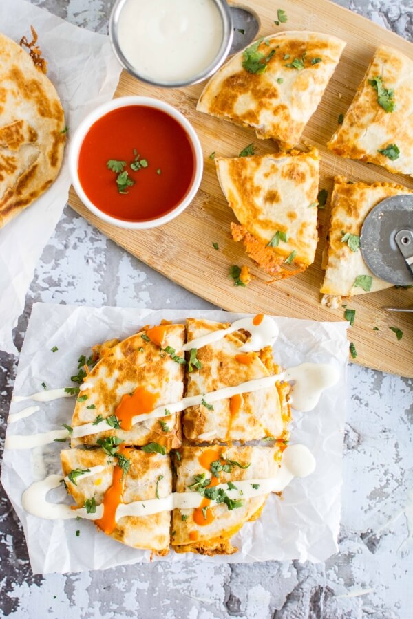 four pieces of quesadilla on parchment paper, 4 pieces of quesadilla on cutting board with 2 small bowls of ranch dressing and red sauce 