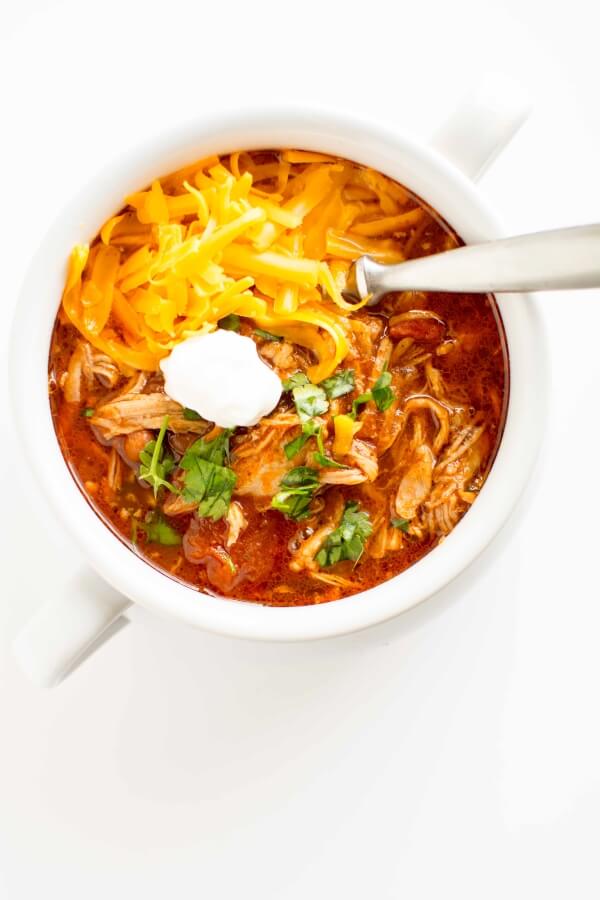 white bowl filled with pulled pork chili, topped with cheese, sour cream and cilantro with spoon