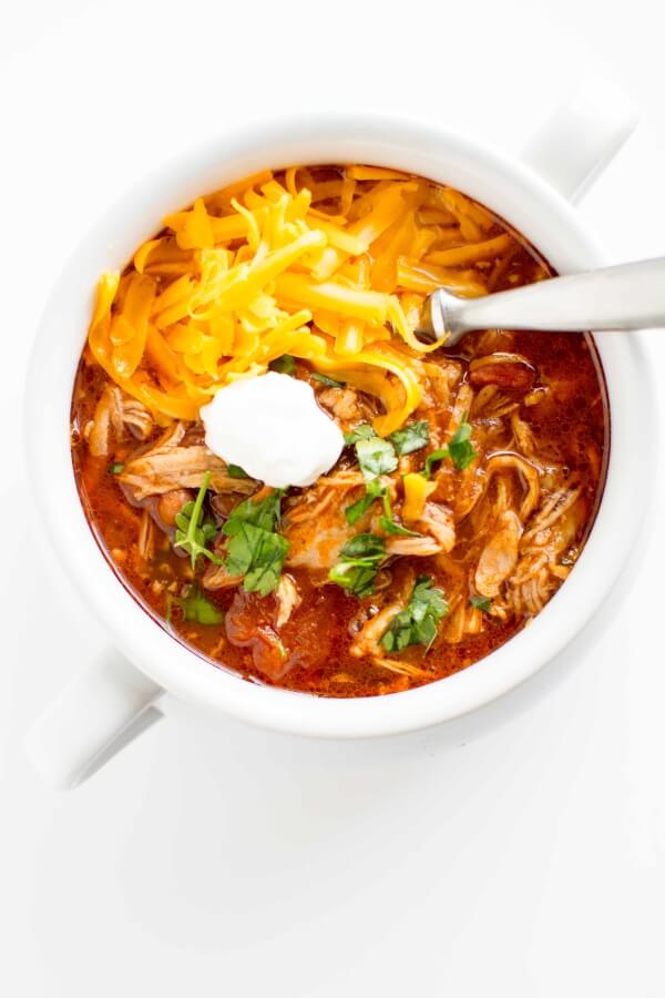 white bowl filled with pulled pork chili, topped with cheese, sour cream and cilantro with spoon
