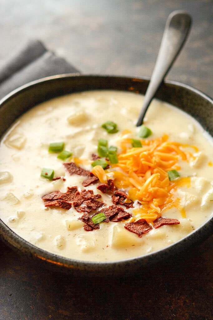 baked potato soup in gray bowl with spoon with bacon, cheese and green onion on top