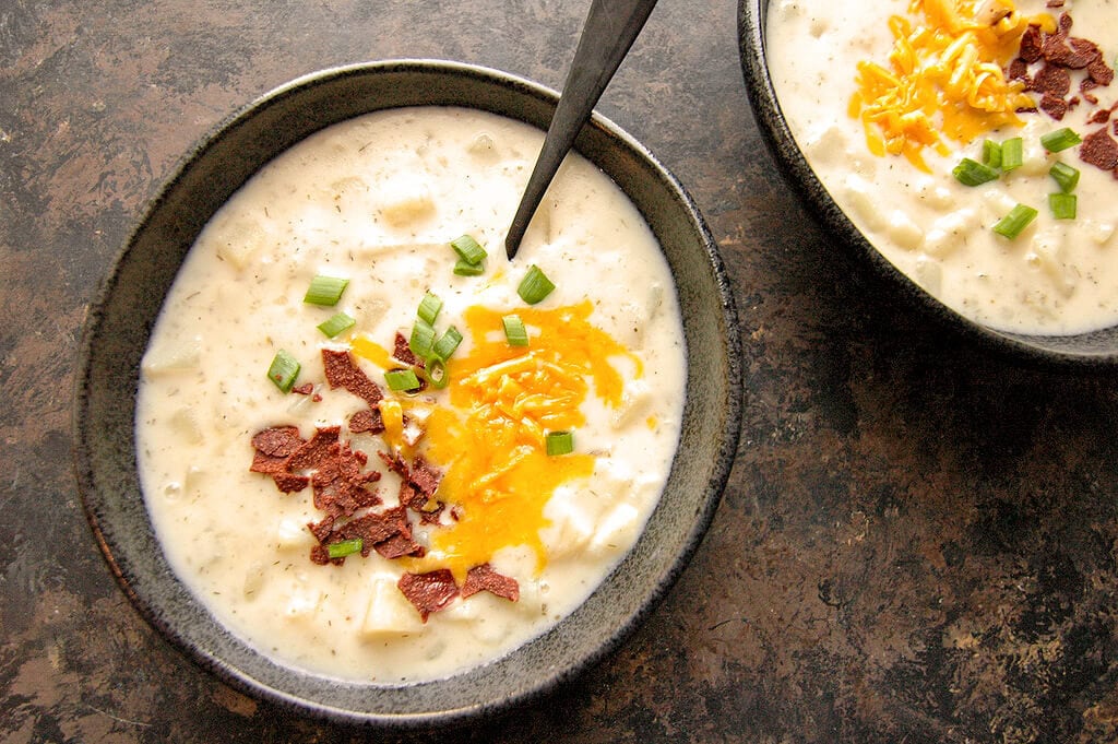 Two servings of Slow Cooker Loaded Baked Potato Soup topped with bacon and cheese in black bowls.