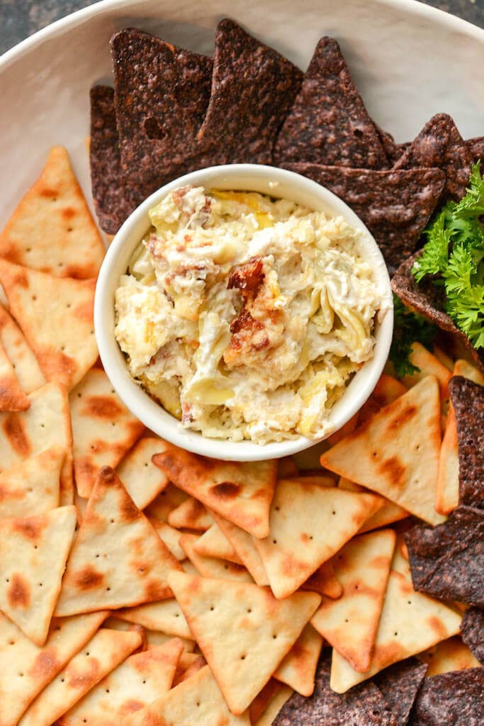 Slow Cooker Artichoke Dip in white bowl with platter of variety of crackers