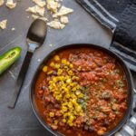 Instant Pot Chili in black bowl on gray table topped with corn