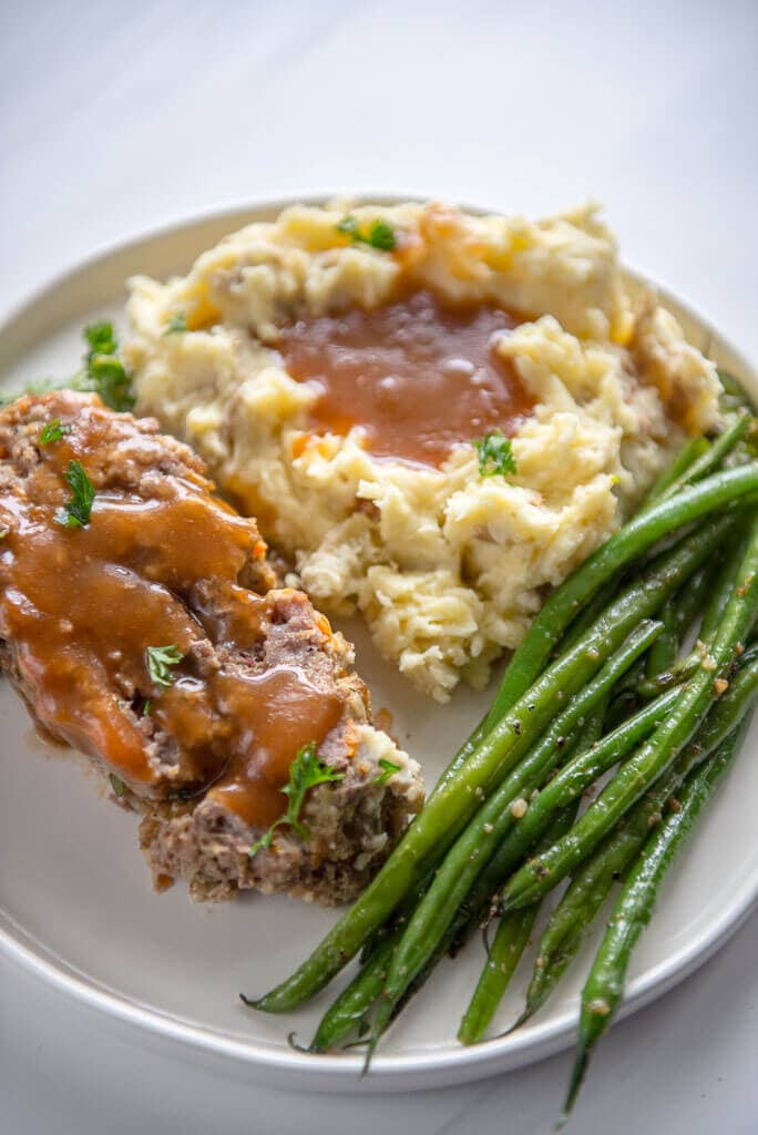 Slow Cooker Meatloaf with Buttermilk Mashed Potatoes on a white plate. Served with crispy garlic onion green beans on the side. 