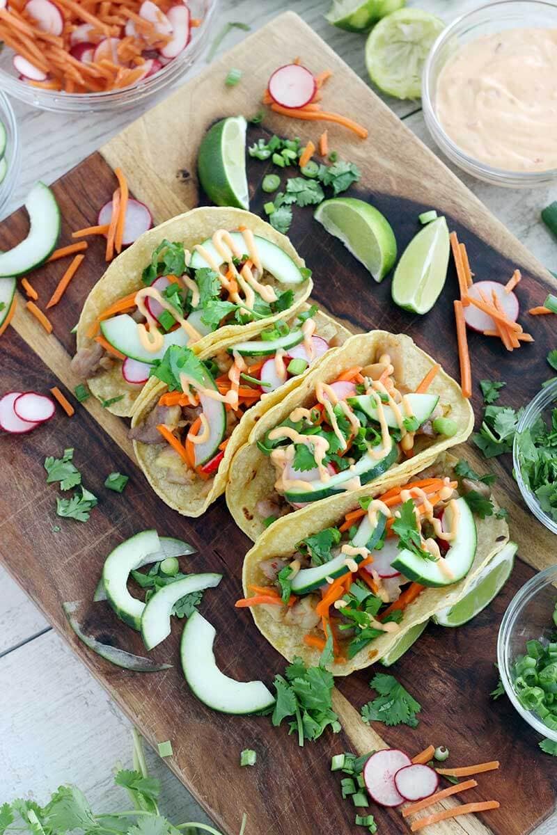 garnished chicken banh mi tacos on wood board with limes and veggies