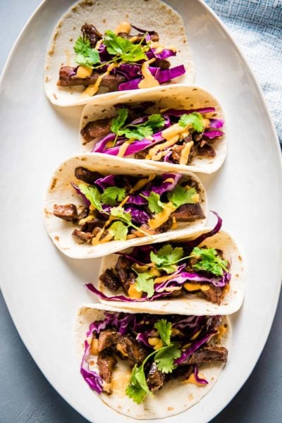 Korean beef tacos on a white platter topped with red cabbage and Sriracha mayo
