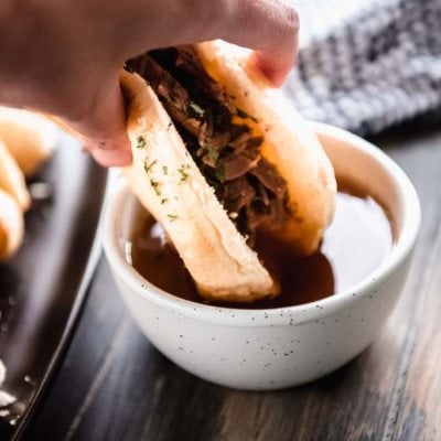 hand holding French dip into au jus sauce in white bowl