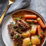 white plate with serving of pot roast, carrots, potatoes and gravy topped with parsley