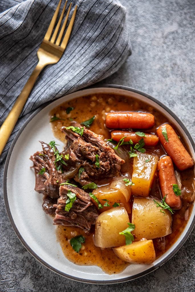 white plate with serving of pot roast, carrots, potatoes and gravy topped with parsley