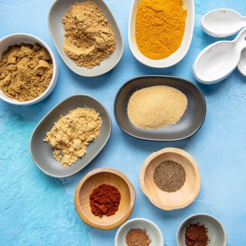 tiny bowls filled with spices to make curry seasoning on a blue backdrop