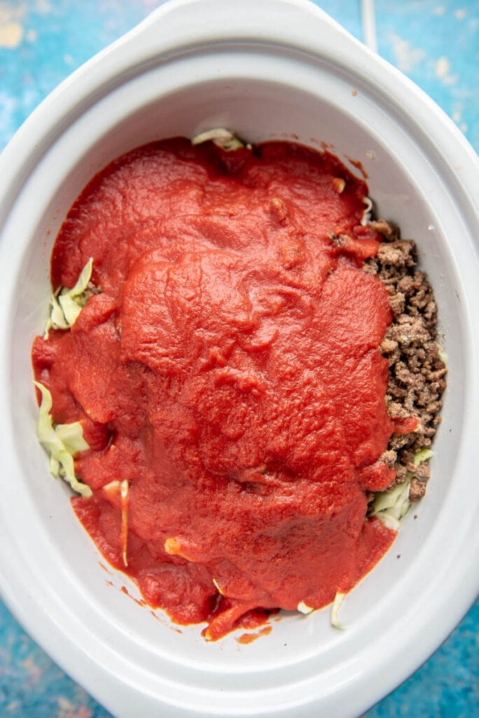 white oval slow cooker filled with beef, cabbage and topped with tomato sauce