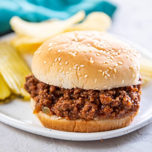 sloppy Joe with bun on white plate with pickles and chips