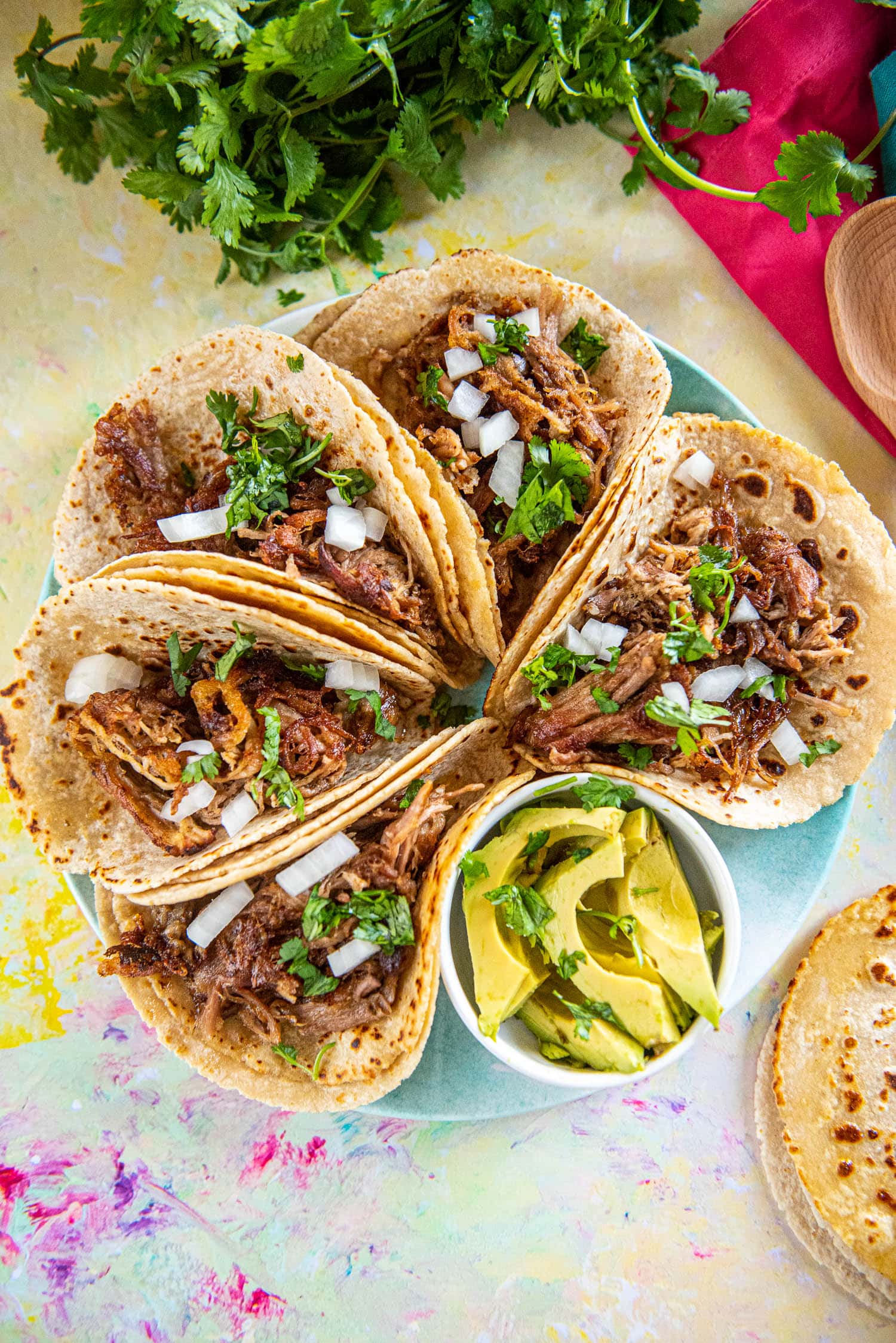 5 street tacos in corn tortillas on round plate with small bowl of sliced avocado