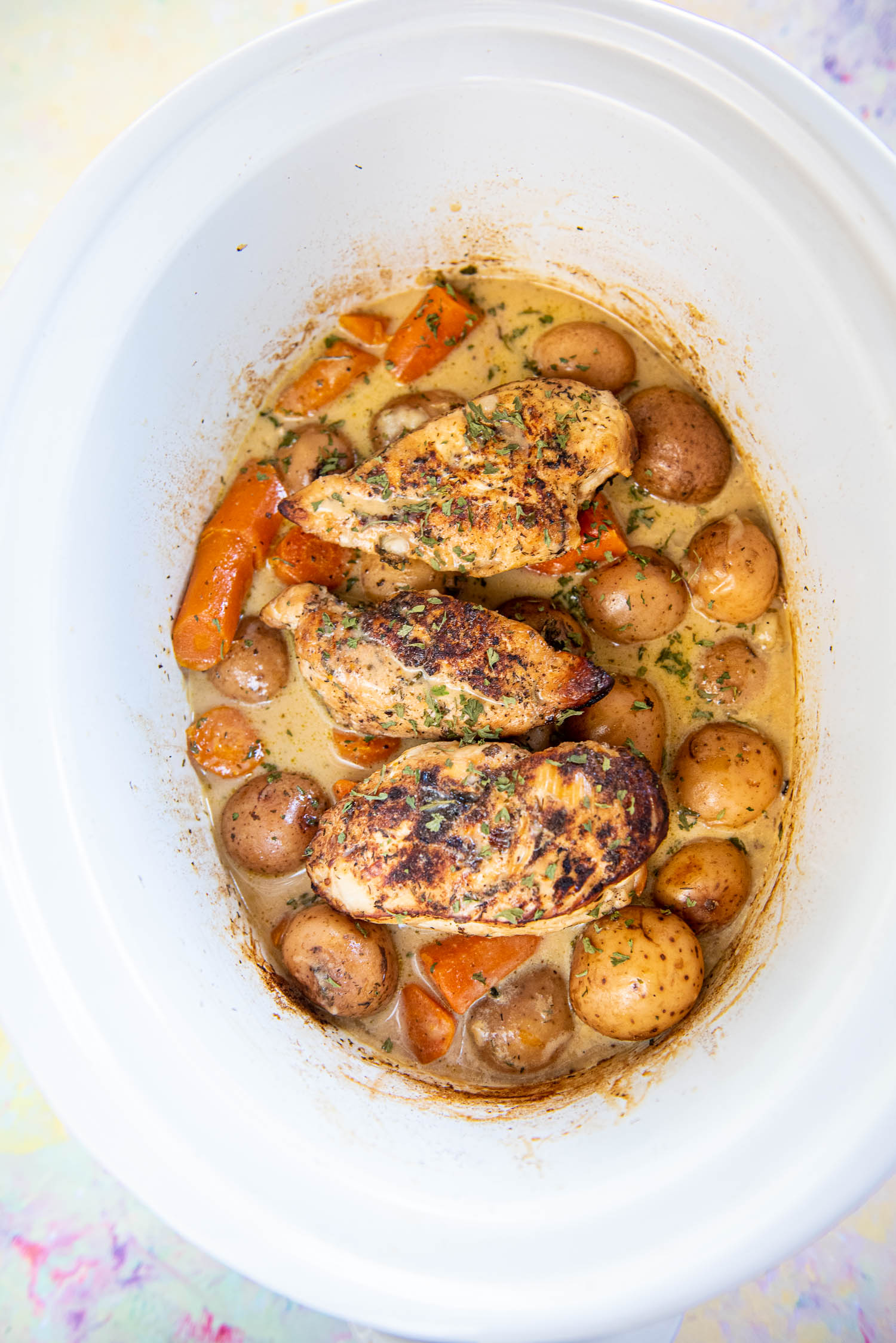 overhead view of white slow cooker with cooked baby potatoes, carrot chunks, chicken breasts and gravy