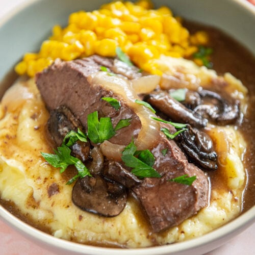 served plate of mashed potatoes, corn niblets, and slow cooker round steak and mushrooms garnished with onions on pink surface