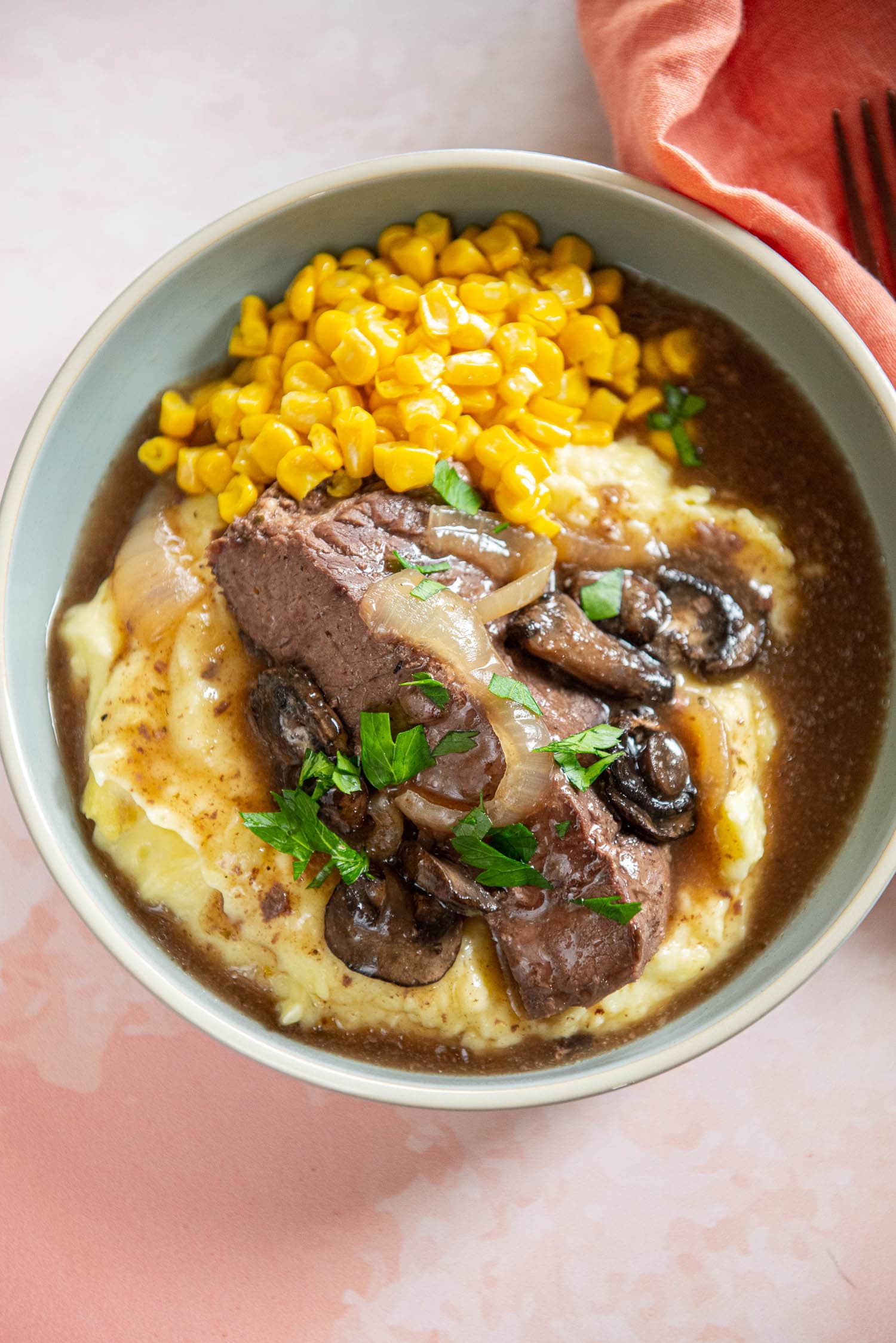 served plate of mashed potatoes, corn niblets, and slow cooker round steak and mushrooms garnished with onions on pink surface