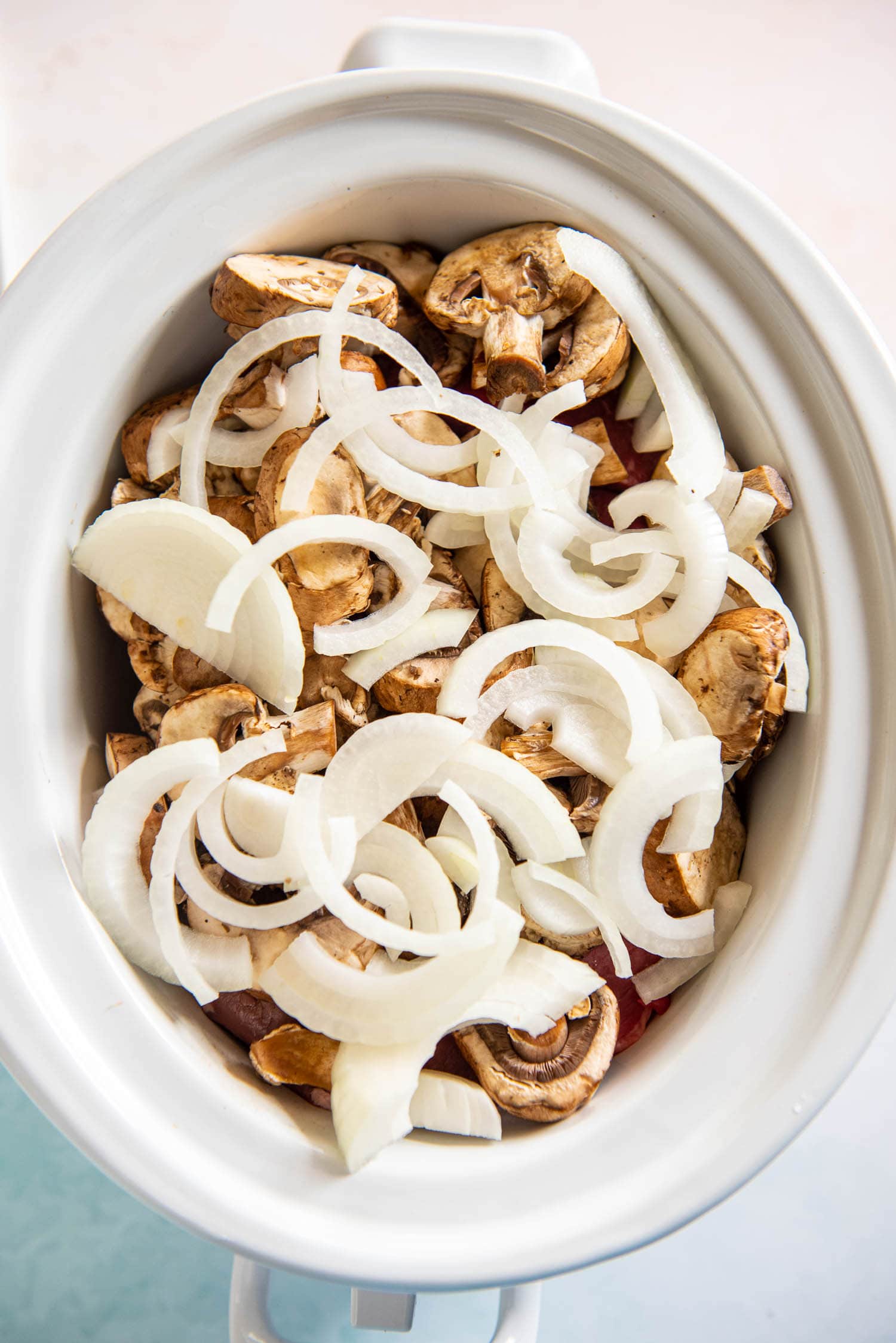 slow cooker containing grass fed angus beef, baby bella mushrooms, and sliced onions on top