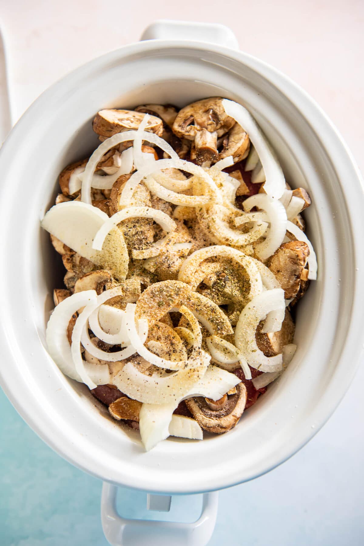 slow cooker containing grass fed angus beef, baby bella mushrooms, and sliced onions and spices on top