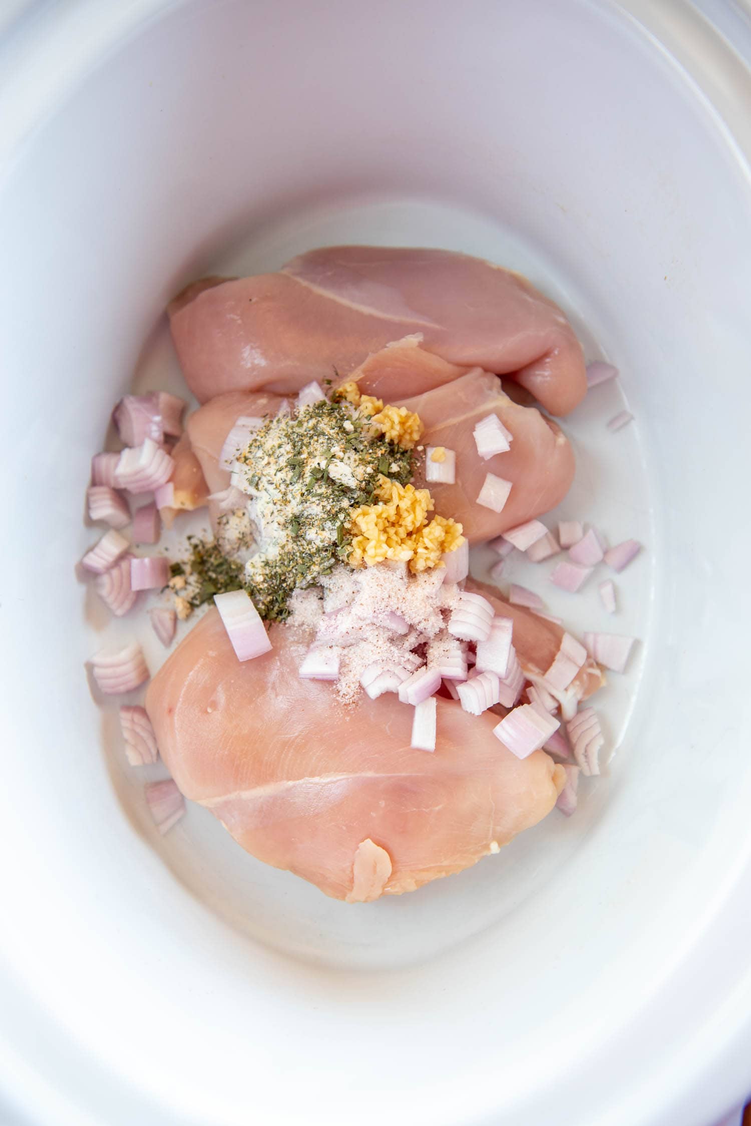 raw chicken breasts inside of white slow cooker down with chopped red onions, small scoop of minced garlic, and green and white spiced on top