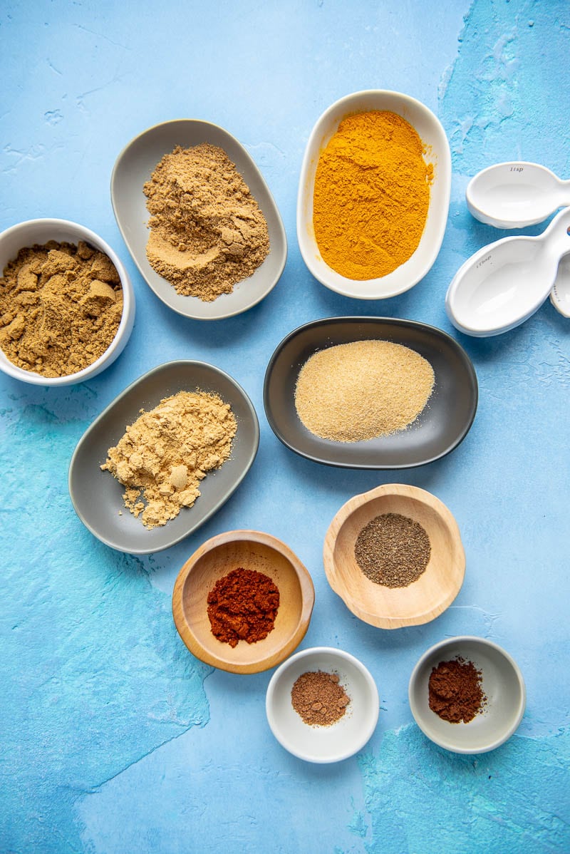 Variety of 9 seasonings and spices in various types and sizes of dishes next to 3 spoons on a blue surface