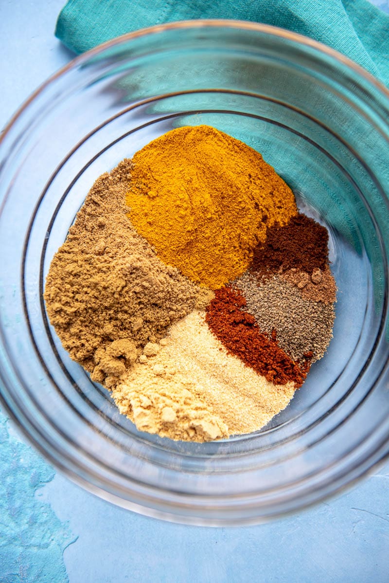 6 spices of different colors like dark red, brown, and burnt orange all shows in a clear glass bowl on top of blue surface