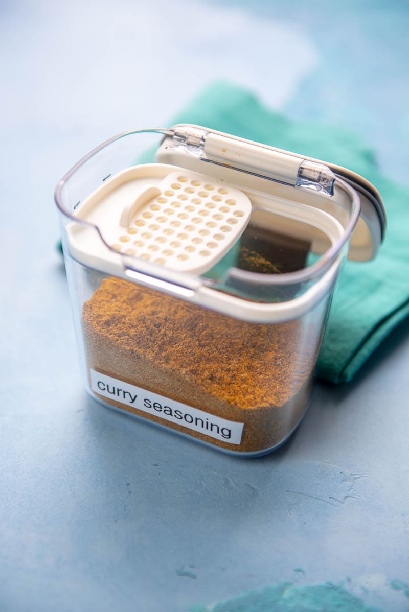 seasoning container labeled "curry seasoning" with curry seasoning in it next to a teal towel on a blue surface