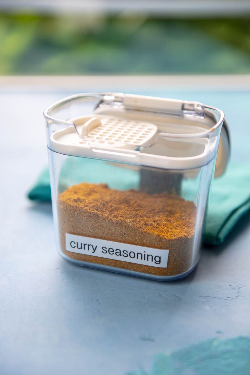 seasoning container labeled "curry seasoning" with curry seasoning in it next to a teal towel on a blue surface