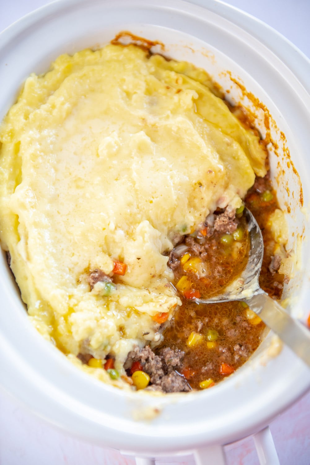 ground beef cooked with with mixed veggies like corn, onions, carrots, and green peas mixing with creamed mashed potatoes on top inside in white slow cooker pot and a large sliver spoon scooping a spoonful of mixture