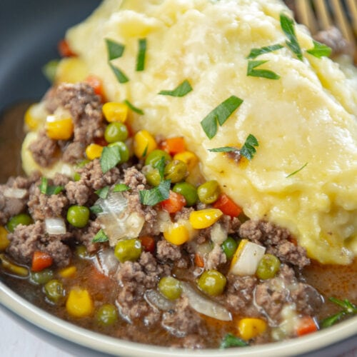 shepherds pie ground beef, corn, peas, carrots, onions, with a side of mashed potatoes garnished on a blue bowl and a gold fork inside