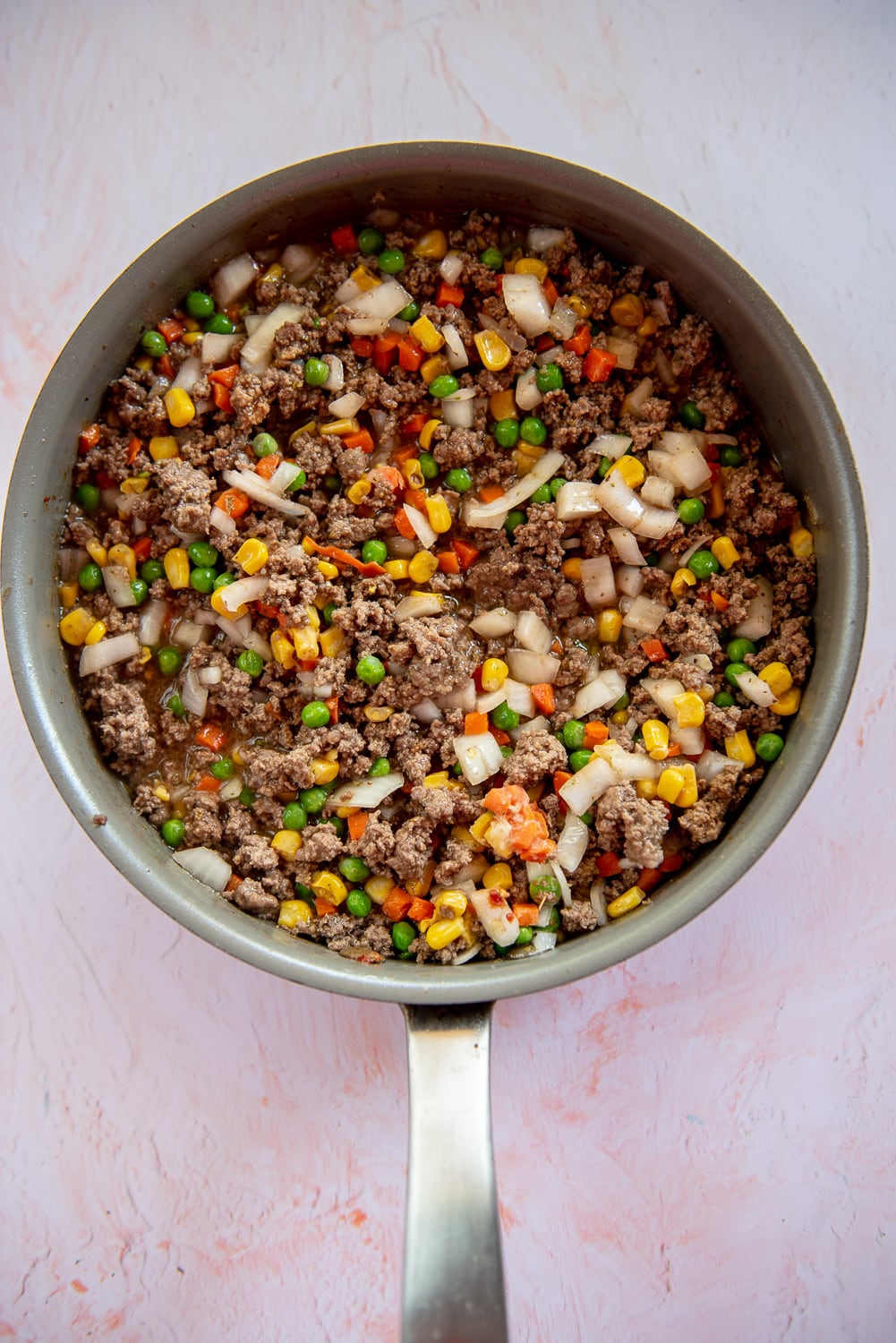ground beef cooked with with mixed veggies like corn, onions, carrots, and green pies inside in silver skillet with pink background