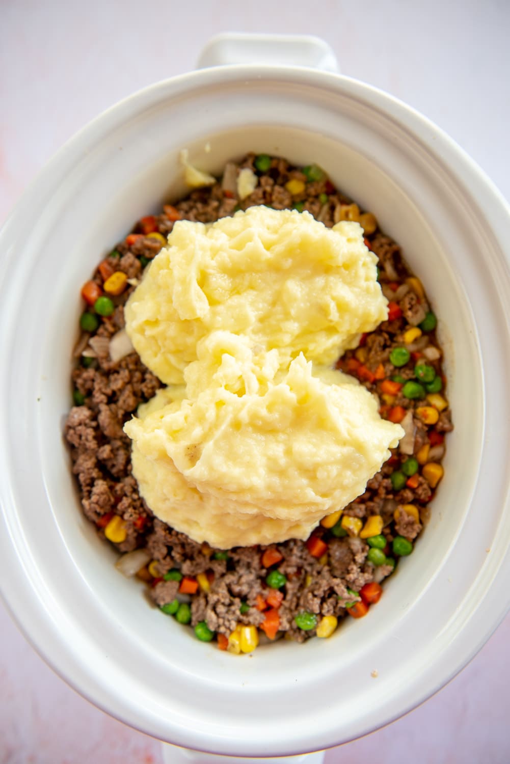 ground beef cooked with with mixed veggies like corn, onions, carrots, and green peas with a dollop of creamed mashed potatoes on top inside in white slow cooker pot